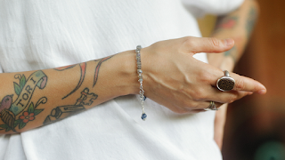 Tattooed hand with a silver beaded bracelet and dangling charm, styled with a bold statement ring and white shirt, showcasing personal fashion expression.